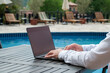 © Evrymmnt - Young man wearing white linen shirt working on his laptop by the pool. Male travel blogger typing on keyboard. Perks of being a freelancer. Close up, copy space, background.