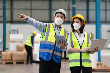 Wall Mural - Asian male supervisor engineer in helmet safety wearing hygienic mask to protect coronavirus with female technician holding laptop point to storage location of products in warehouse factory industrial