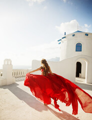 Fashion outdoor photo of gorgeous young woman with red hair in airy red dress posing on the streets of Oia, Santorini, Greece. 