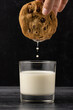 Hand dipping chocolate chip cookie in glass of milk against dark background
