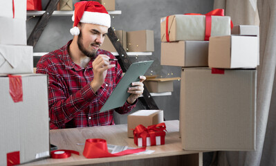 Portrait of a bearded guy Santa Claus in a hat works in a warehouse of an online store prepares goods in boxes for shipment for New Year and Christmas. New 2022 gifts for children.