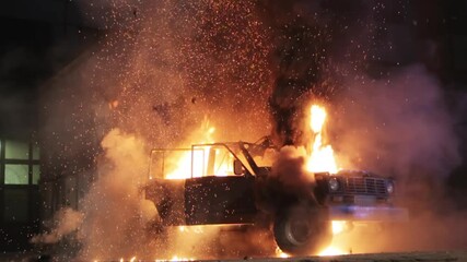 The car explodes and the hood and roof are torn off. A black SUV is torn apart by a huge orange flame of an explosion, the night on a city street. The car explodes from a bomb or a shell hit.