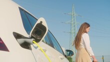 Handheld shot of a young Caucasian woman leaning against the еlectric car with the charger plugged in, checking her smartphone, in the background visible transmission line