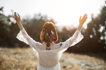 Indian boho vibes, bohemian style. Back view of stunning hippie boho woman with tattoos, wearing feathers in short ginger hair, white dress, stands with arms raised and outstretched to sunset in field