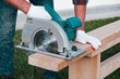 Working with a power tool. A builder is sawing a board at the construction site of a new house. Close-up on gloved hands holding a circular saw.