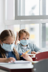 Wall Mural - schoolboy in medical mask raising hand during lesson near blurred girl