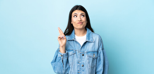 Wall Mural - Young brunette girl over isolated blue background with fingers crossing and wishing the best