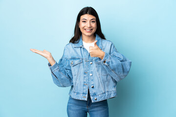 Young brunette girl over isolated blue background holding copyspace imaginary on the palm to insert an ad and with thumbs up