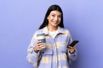 Wall Mural - Young brunette woman over isolated purple background holding coffee to take away and a mobile