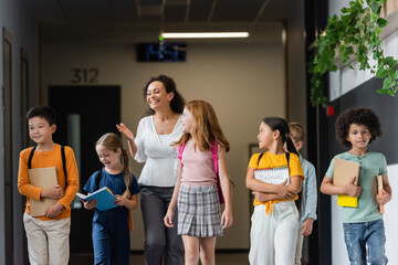 Wall Mural - smiling african american teacher pointing with hand while talking to multiethnic children in school hall