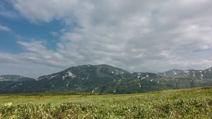 Wall Mural - The meadow is covered with lush grass. A mountain range against a background of blue sky and clouds. There is green vegetation on the slopes, areas of snow. Kamchatka