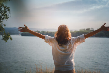 Wall Mural - Happy beautiful young woman enjoying the view and landscape on the river bank with hands raised up at sunset