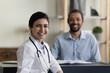 © fizkes - Portrait of smiling confident young female indian ethnicity gp doctor physician posing in clinic with blurred african american male patient on background, professional medicare healthcare services.
