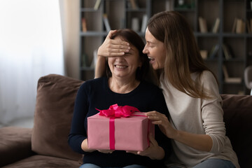 Canvas Print - Happy millennial beautiful woman covering eyes of happy middle aged elderly mother, presenting wrapped surprise gift, congratulating with international woman's day or celebrating happy birthday.