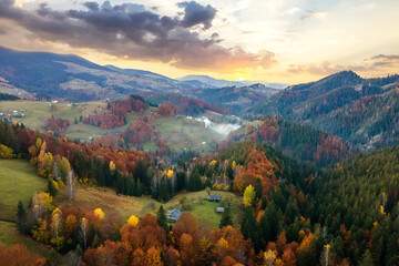 Wall Mural - Aerial view of distant village with small shepherd houses on wide hill meadows between autumn forest trees in Ukrainian Carpathian mountains at sunset.