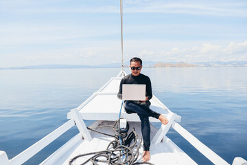 Asian man in wetsuit and sunglasses sitting on the boat and using a laptop on vacation at Labuan Bajo