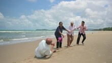 Happy Asian Elderly Tourist Playing Tug Of War Together And Fun On Beach In Outdoors. Group Of Senior Retirement And Friends Relax By Pulling Rope During Tug Of War Game At Coast In Summer Vacation. 