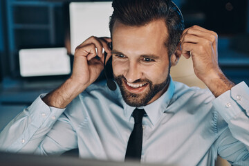 Canvas Print - Happy young man in headset looking at the computer and smiling while staying late in the office