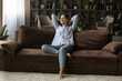 © fizkes - Full length smiling woman relaxing on couch at home, happy dreamy beautiful young female leaning back with hands behind head, visualizing good future, new opportunities, enjoying weekend