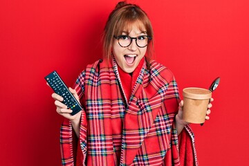 Wall Mural - Redhead young woman wrapped in a red warm red blanket eating icecream and watching tv celebrating crazy and amazed for success with open eyes screaming excited.