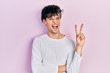 Handsome hipster young man wearing casual white shirt smiling with happy face winking at the camera doing victory sign. number two.