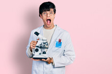 Handsome Hipster Young Man Wearing Lab Coat Holding Microscope Afraid And Shocked With Surprise And Amazed Expression, Fear And Excited Face.