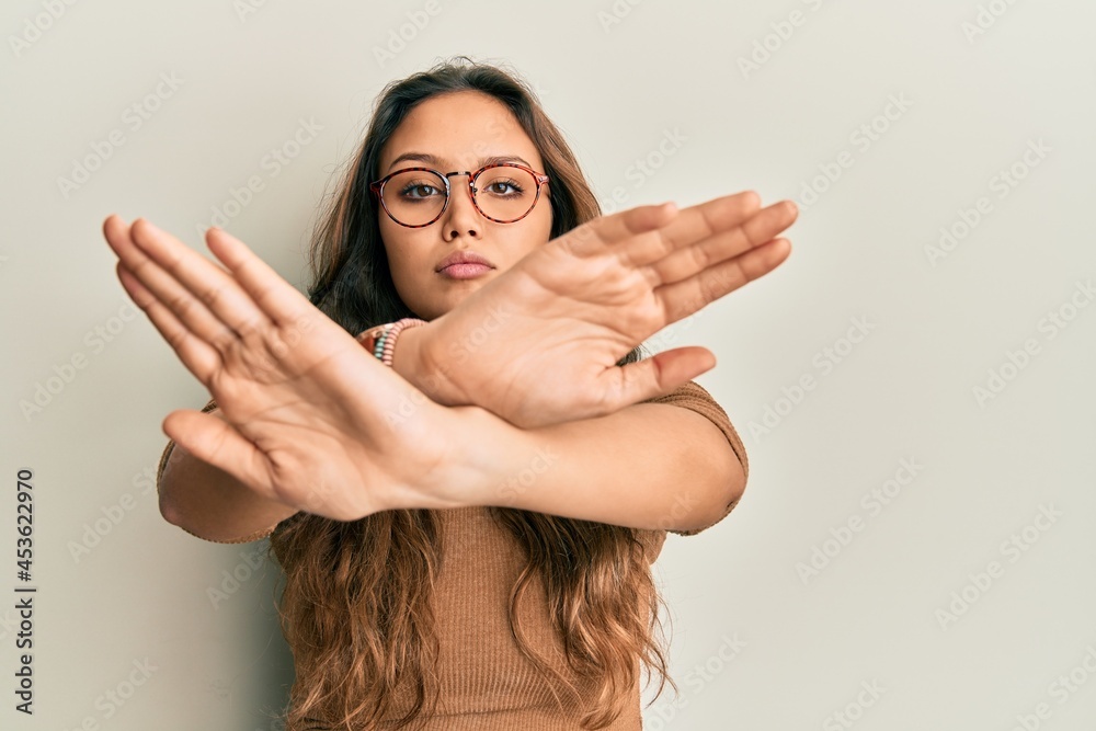 Young hispanic girl wearing casual clothes and glasses rejection expression crossing arms and palms doing negative sign, angry face