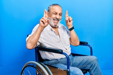 Poster - Handsome senior man with beard sitting on wheelchair gesturing finger crossed smiling with hope and eyes closed. luck and superstitious concept.