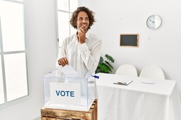 Sticker - Young hispanic man voting putting envelop in ballot box with hand on chin thinking about question, pensive expression. smiling and thoughtful face. doubt concept.