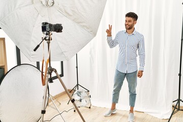 Sticker - Arab young man posing as model at photography studio smiling looking to the camera showing fingers doing victory sign. number two.