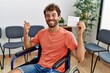 Young handsome man holding covid record card sitting on wheelchair pointing thumb up to the side smiling happy with open mouth