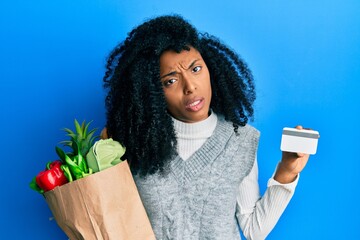 Canvas Print - African american woman with afro hair holding groceries and credit card clueless and confused expression. doubt concept.