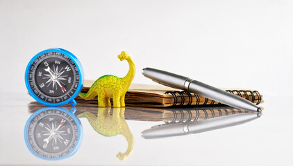 Compass, fountain pen and notepad next to a funny toy dinosaur - diplodocus. White reflective background. Vacation planning concept, travel or tourist trip