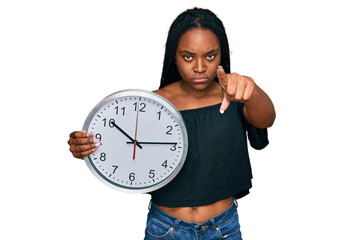 Sticker - Young african american woman holding big clock pointing with finger to the camera and to you, confident gesture looking serious