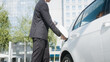 canvas print picture - Businessman plugging in the charger in electric car and going to work in business center - skyscraper