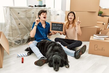 Poster - Young caucasian couple with dog holding our first home blackboard at new house pointing finger up with successful idea. exited and happy. number one.