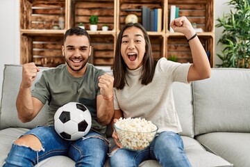 Canvas Print - Young latin couple watching soccer match eating porpcorn at home.