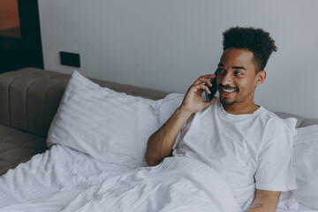 Wall Mural - Young happy satisfied african american man in nightwear speak mobile cell phone lying in bed rest relax spend time in bedroom lounge home in own room house wake up dream be lost in reverie good day.