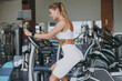 Side view minded young strong sporty athletic sportswoman woman 20s wearing white sportswear earphones listen music warm up training use exercise bike in gym indoor Workout sport motivation concept.