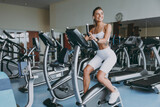 Full body young skinny strong sporty athletic sportswoman caucasian woman in white sportswear earphones listen music look aside warm up train use exercise bike in gym indoors. Workout sport concept.