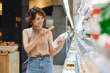 Wall Mural - Young woman in casual clothes backpack shopping at supermaket store buy dairy produce take milk take photo by mobile cell phone of ingredients inside hypermarket Purchasing gastronomy food concept