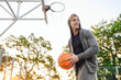 Young bottom view excited smiing sporty sportsman man in grey sports clothes hood look aside training holding in hand ball play at basketball game playground court. Outdoor courtyard sport concept