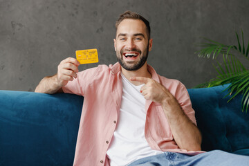 Wall Mural - Young smiling happy fun man 20s in casual clothes point index finger on credit bank card sitting on blue sofa at home flat indoors rest relax on weekends free time. People lounge lifestyle concept