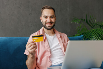 Poster - Young satisfied fun man in casual clothes using work on laptop pc computer credit bank card shopping online order delivery booking tour sitting on blue sofa at home flat indoor rest relax on weekends