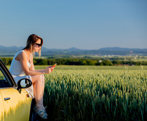 Canvas Print - young girl in sunglasses with smartphone is sitting on a car on green wheat field in spring time