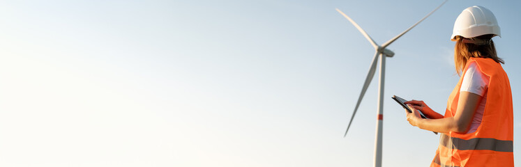 An engineer looks at a wind turbine and holds a tablet in his hands on the banner with copy space. 