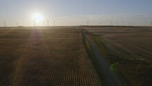 Aerial Flying Over A Road In North Dakota Farmland Towards Windmills In The Evening With The Sun Setting And Distant Windmills Spinning