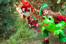 Santa Claus With Elves On The Background Of The Forest, A Funny Girl In An Inflatable Circle Of Toads, Santa Is Talking While Sitting In A Chemonade With Bright Toys For Christmas And New Year