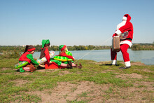 Santa Claus Led Elf Girls On A Sled Against The Background Of The River In The Summer In Inflatable Circles Hurries For Christmas And New Year's Eve For The Holiday. Hotel Holiday Concept