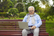© Studio Romantic - An elderly happy man of Asian appearance sits on a bench in a proud park with a laptop. Senior man gray-haired video communication with grandchildren, distance communication concept, covid 19